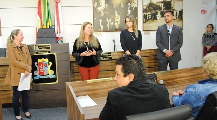 na câmara de vereadores, duas mulheres em pé falam; vereadora meri de um lado observa; e de outro um homem de terno. algumas pessoas sentadas na frente