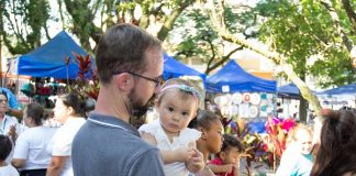 menina bebê no colo de um homem olha para a câmera; ao redor pessoas circulam e barracas da feira ao fundo, com algumas árvores