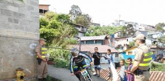 piloto faz uma curva de um caminho do morro observado por dois pm e um grupo de crianças ao lado