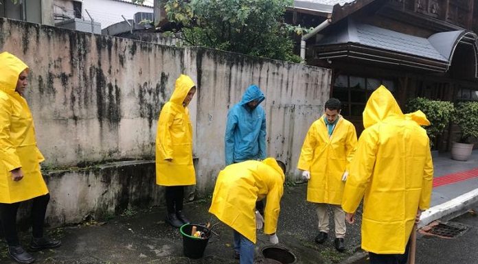 em um dia nublado, homens com capas de chuva amarelas estão em pé parados em volta de um bueiro olhando para o chão