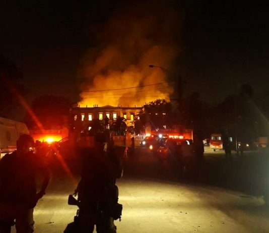 foto noturna da fachada do prédio histórico em chamas; algumas silhuetas de agentes de segurança na frente