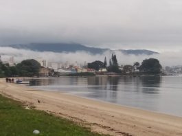 praia com mar calmo e enseada ao fundo com nevoeiro sobre o continente