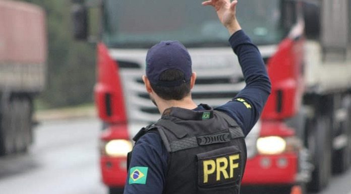 agente da prf visto de costas com uniforme da corporação gesticula com o braço direito para caminhão na estrada