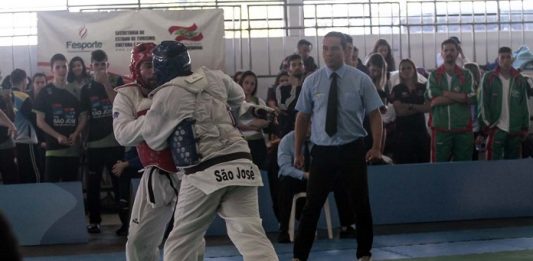 dois competidores de taekwondo lutam, um com capacete azul, outro vermelho, observados pelo juiz no tatame e público em geral na arquibancada em volta