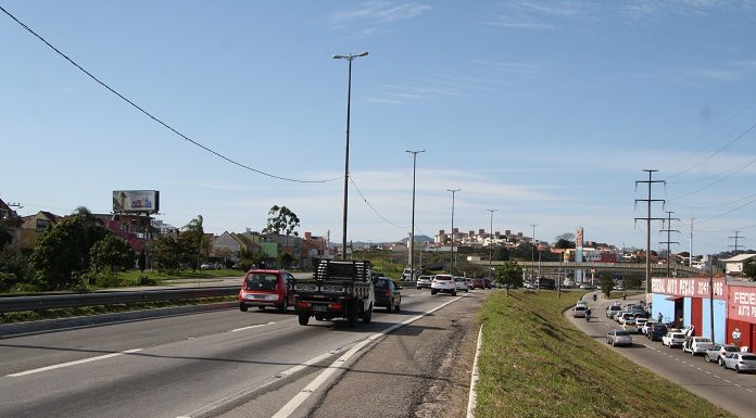 carros passam pela rodovia em foto tirada no acostamento em dia ensolarado