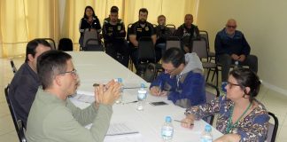 em uma mesa onde estão duas pessoas de cada lado, homem gesticula conversando com uma mulher à sua frente; ao fundo da sala, pessoas assistem sentadas em cadeiras lado a lado