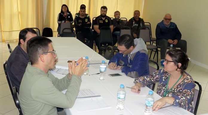 em uma mesa onde estão duas pessoas de cada lado, homem gesticula conversando com uma mulher à sua frente; ao fundo da sala, pessoas assistem sentadas em cadeiras lado a lado