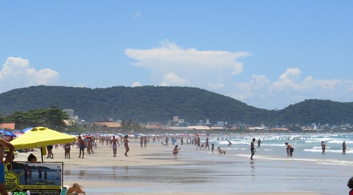 praia larga em um dia ensolarado; pessoas são vistas na distância circulando; algumas em primeiro plano debaixo de guarda sol