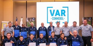 arbitros posam com certificados em saguão de hotel com uma grande placa ao fundo escrita "var conmebol"