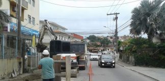 pequena rua de mão dupla, em que uma via está interditada onde há vala aberta por retroescavadeira e operario observa