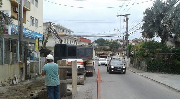 pequena rua de mão dupla, em que uma via está interditada onde há vala aberta por retroescavadeira e operario observa