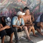 homens moradores de rua sentados em pequena sarjeta junto a muro grafitado olham na mesma direção