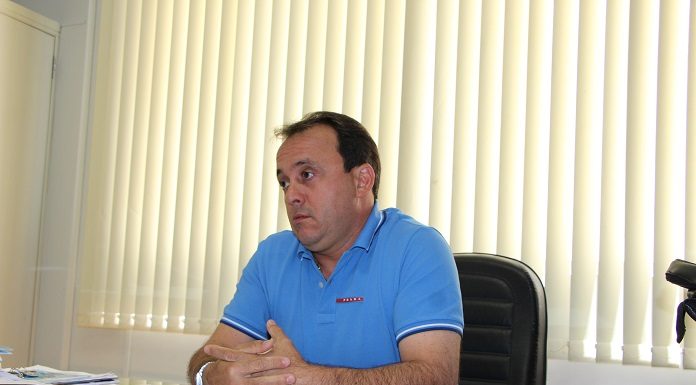 juliano em seu gabinete sentado à frente de uma mesa cheia de papeis, com uma persiana fechada ao fundo