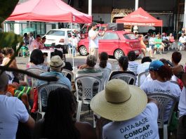 trabalhadores com camisas escritas "dasta-base" sentados assistem a um homem discursas; carro e tenda ao fundo do sindicato