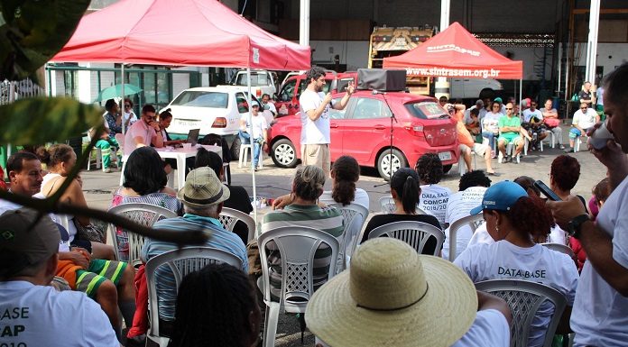 trabalhadores com camisas escritas "dasta-base" sentados assistem a um homem discursas; carro e tenda ao fundo do sindicato