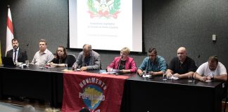 diversas pessoas em uma bancada à frente de um auditório; há uma bandeira do movimento de população de rua sobre a bancada; deputada carminatti ao centro fala no microfone