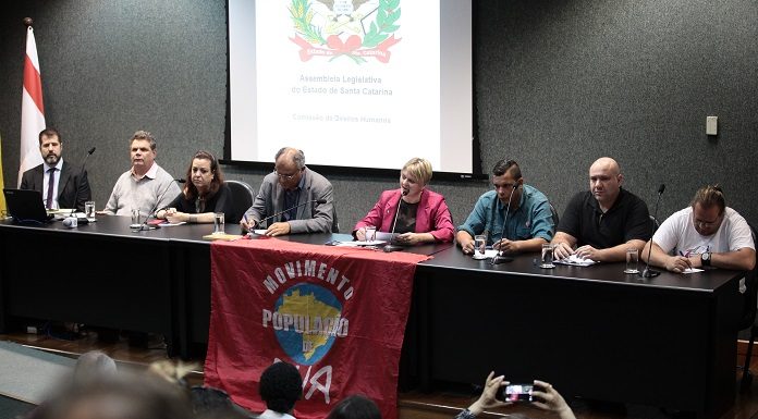 diversas pessoas em uma bancada à frente de um auditório; há uma bandeira do movimento de população de rua sobre a bancada; deputada carminatti ao centro fala no microfone