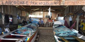 dois barcos guardados dentro de um rancho de pesca, com chão de areia; algumas pessoas ao fundo do rancho