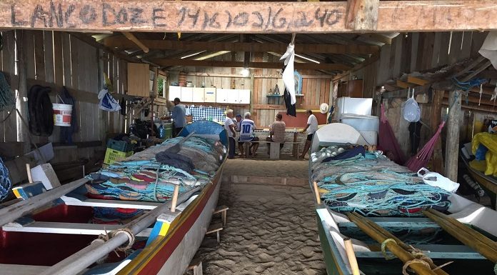 dois barcos guardados dentro de um rancho de pesca, com chão de areia; algumas pessoas ao fundo do rancho