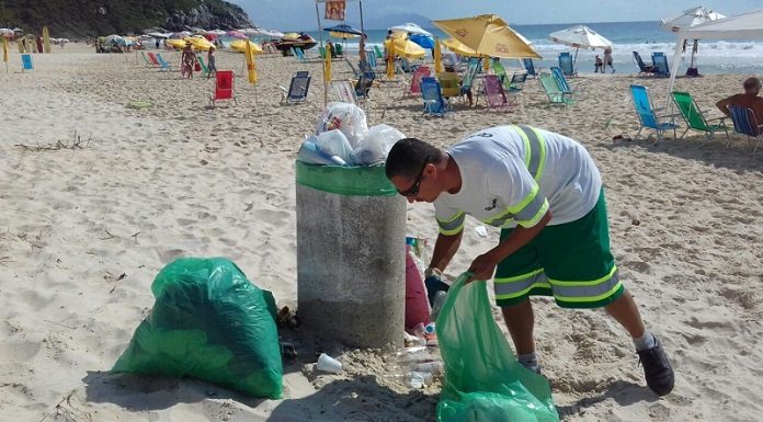 trabalhador da comcap se abaixa para pegar lixo com sacola na mão ao lado de lixeira em uma praia