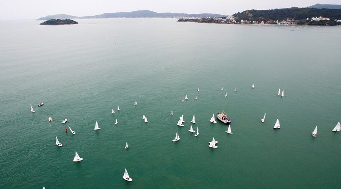 imagem aérrea do mar esverdeado com diversos barcos à vela espalhados; ao fundo algumas montanhas da ilha de sc