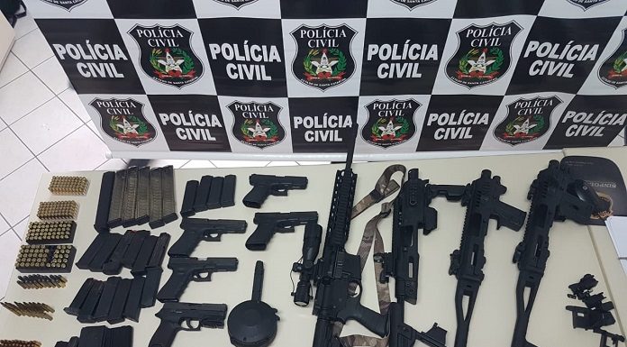 mesa cheia de armas, metralhadoras e pentes de munição à frente de um painel com logos da polícia civil