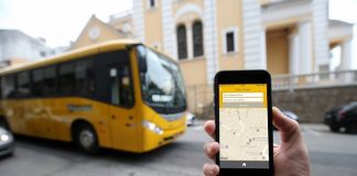 mão em frente à câmera segurando celular com aplicativo aberto e um ônibus amarelo passando atrás