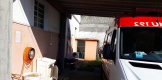 ambulância estacionada em garagem precária junto à uma casa, com máquina de lavar roupa antiga e outros objetos; há um homem sentado ao fundo na sombra