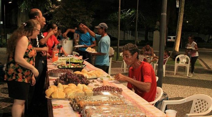 grande mesa com comidas e voluntários ao redor; um homem sentado à mesa comendo