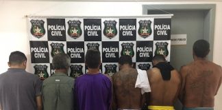 seis homens em pé fotografados de costas, alguns sem camisa, em uma delegacia em frente a um painel com logos da polícia civil