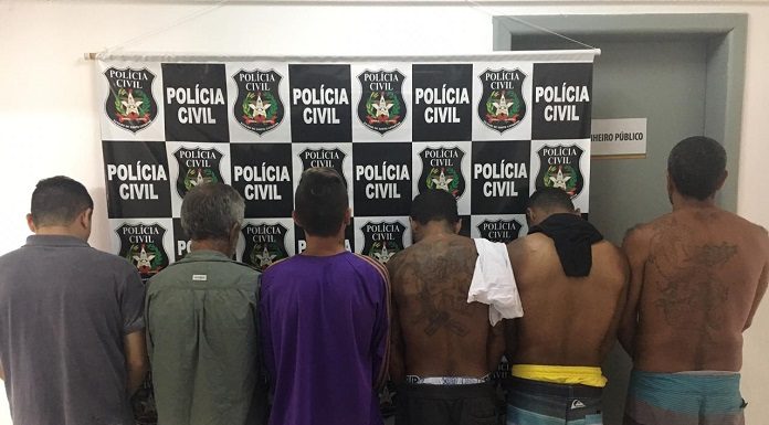 seis homens em pé fotografados de costas, alguns sem camisa, em uma delegacia em frente a um painel com logos da polícia civil