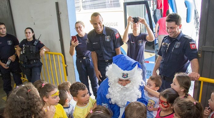 papai noel na cor azul agachado e rodeado de crianças observado por agentes sorridentes da guarda municipal