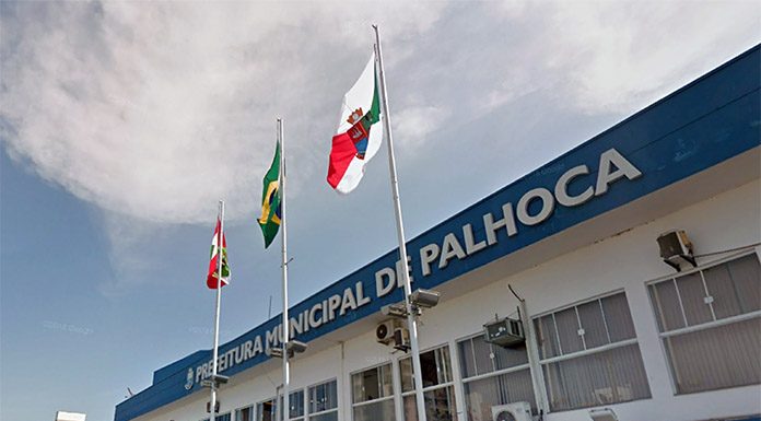 fachada do prédio atual da prefeitura de palhoça com as bandeira de sc, br e do município à frente