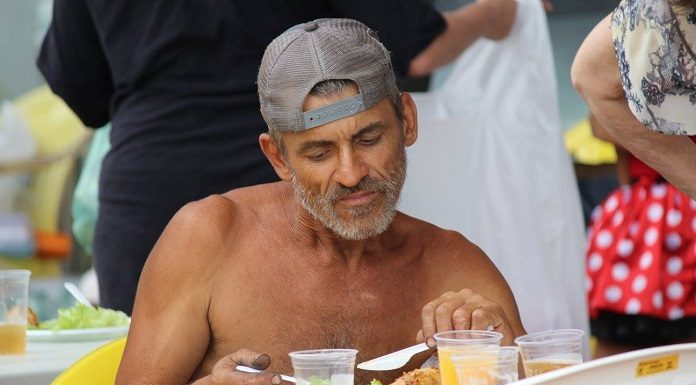 senhor sem camisa e com boné sentado comendo em uma mesa de plástico com alguns copos de refrigerante na frente