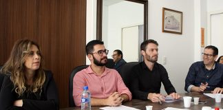 vereadores sentados do mesmo lado de uma grande mesa olham para fora da foto; lemos está sorrindo; há copos de café e garrafa dágua em cima da mesa