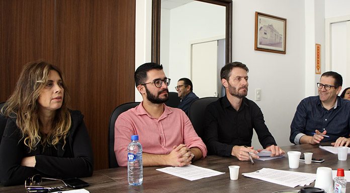 vereadores sentados do mesmo lado de uma grande mesa olham para fora da foto; lemos está sorrindo; há copos de café e garrafa dágua em cima da mesa