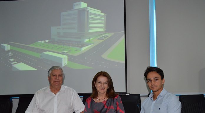 as três pessoas sentadas em uma mesa posam para a câmera com uma projeção da nova sede ao fundo