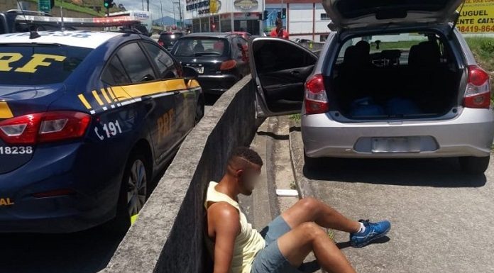 homem sentado no chão e algemado encostado no guard rail ao lado do carro furtado aberto e de viatura da prf