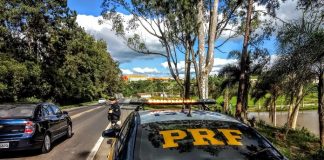 policial rodoviária parada em pé no acostamento na frente de uma viatura olhando carro passar e a estrada