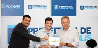 três pessoas, inluindo o prefeito gean, posam para a câmera com o documento em mãos; atrás, um painel com logos da prefeitura de florianópolis