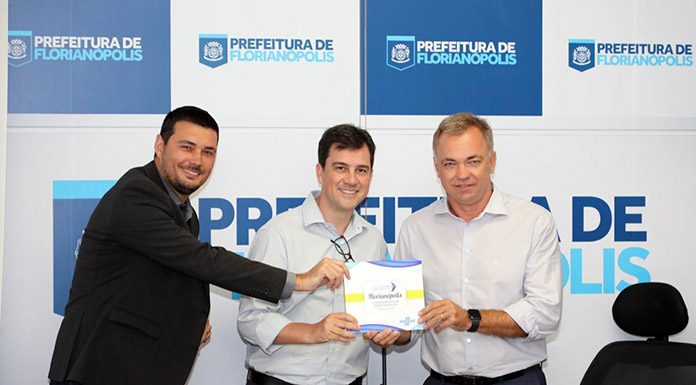 três pessoas, inluindo o prefeito gean, posam para a câmera com o documento em mãos; atrás, um painel com logos da prefeitura de florianópolis