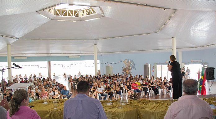 prefeita adeliana vista por detrás do palco onde há pessoas sentadas falando com a grande plateia ao fundo