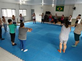 em uma grande sala com tatame no chão, grupo de mulheres faz alongamento coletivo em uma grande roda