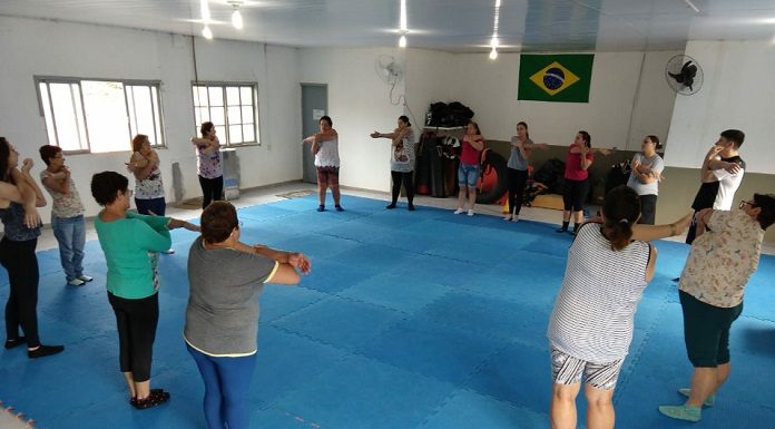 em uma grande sala com tatame no chão, grupo de mulheres faz alongamento coletivo em uma grande roda