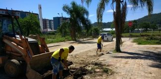 homem tira terra da rua com uma pá ao lado de retroescavadeira; mais à frente outro homem também faz o mesmo movimento; rua de lajotas