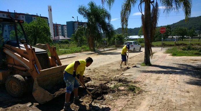 homem tira terra da rua com uma pá ao lado de retroescavadeira; mais à frente outro homem também faz o mesmo movimento; rua de lajotas