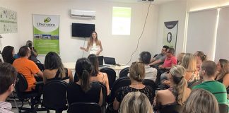 em uma sala lotada de pessoas sentadas, mulher dá palestra ao lado de um banner do ossj