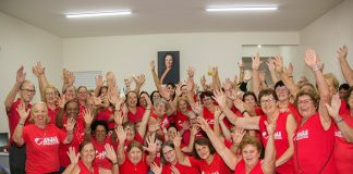 grande grupo de voluntárias usando a camiseta vermelha da associação posa de braços para o alto para a foto; a maioria idosas