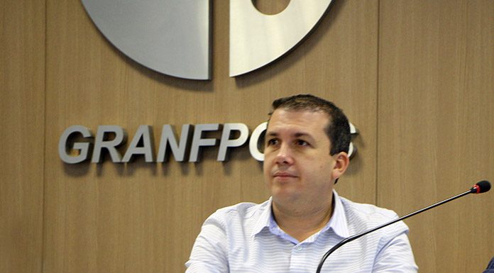 camilo martins sentado em mesa com microfone e escrito granfpolis na parede ao fundo