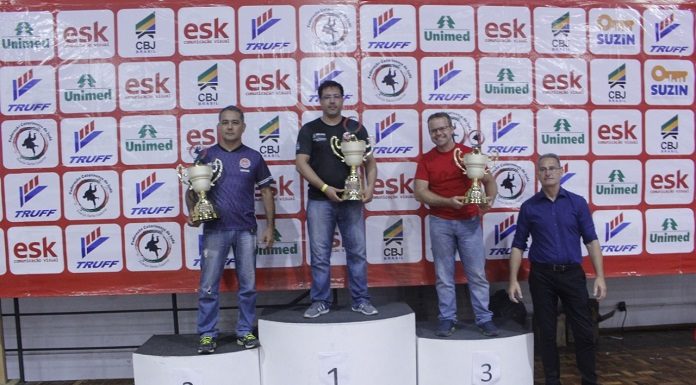 homens no podio segurando trofeus de primeiro, segundo e terceiro lugar em frente a um painel com patrocinadores